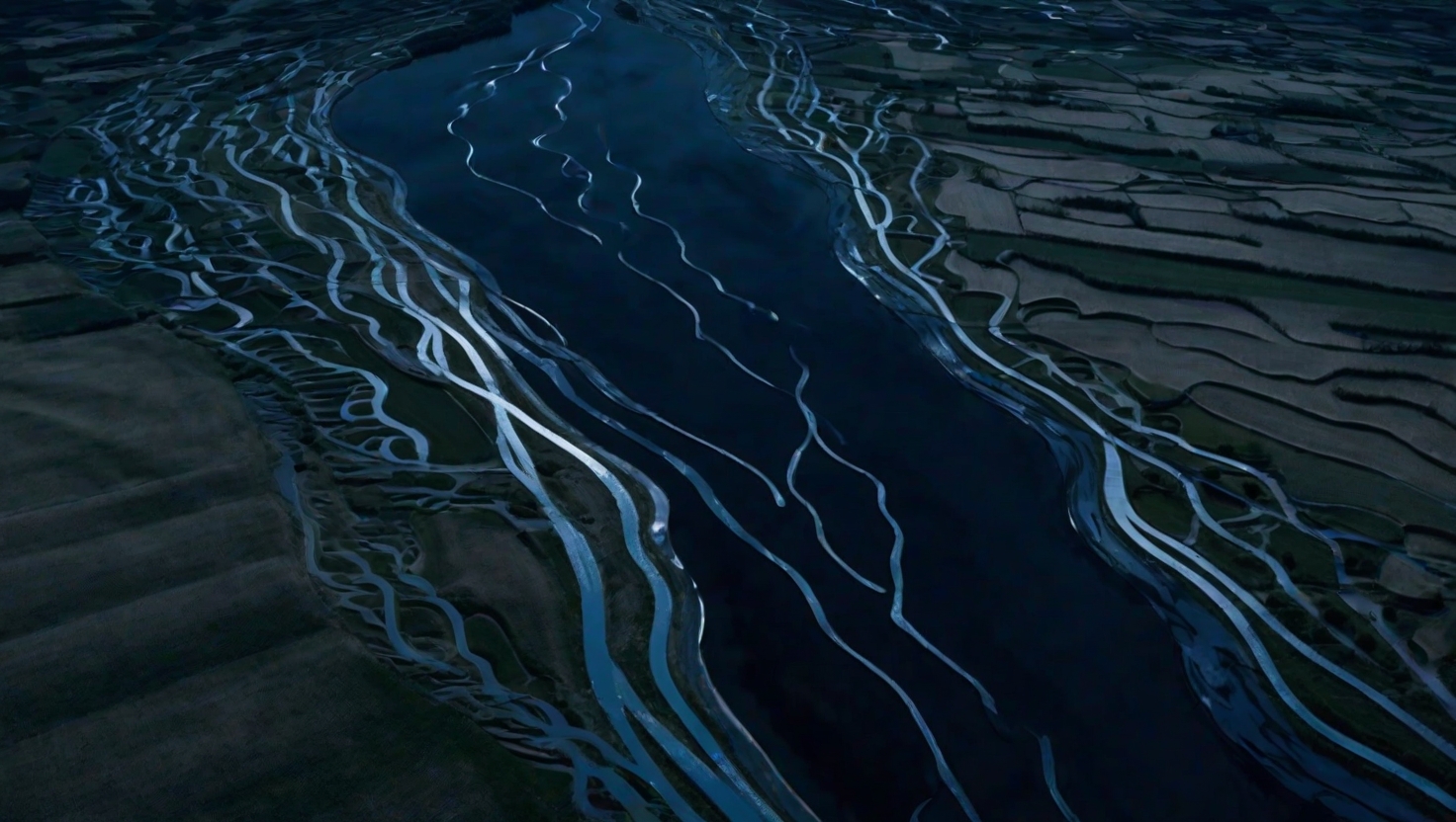 Wide aerial view of a winding river cutting silver ribbons through dark landscape under moonlight, geometric farmland patterns contrasting with the river's organic curves, deep blue and silver tones